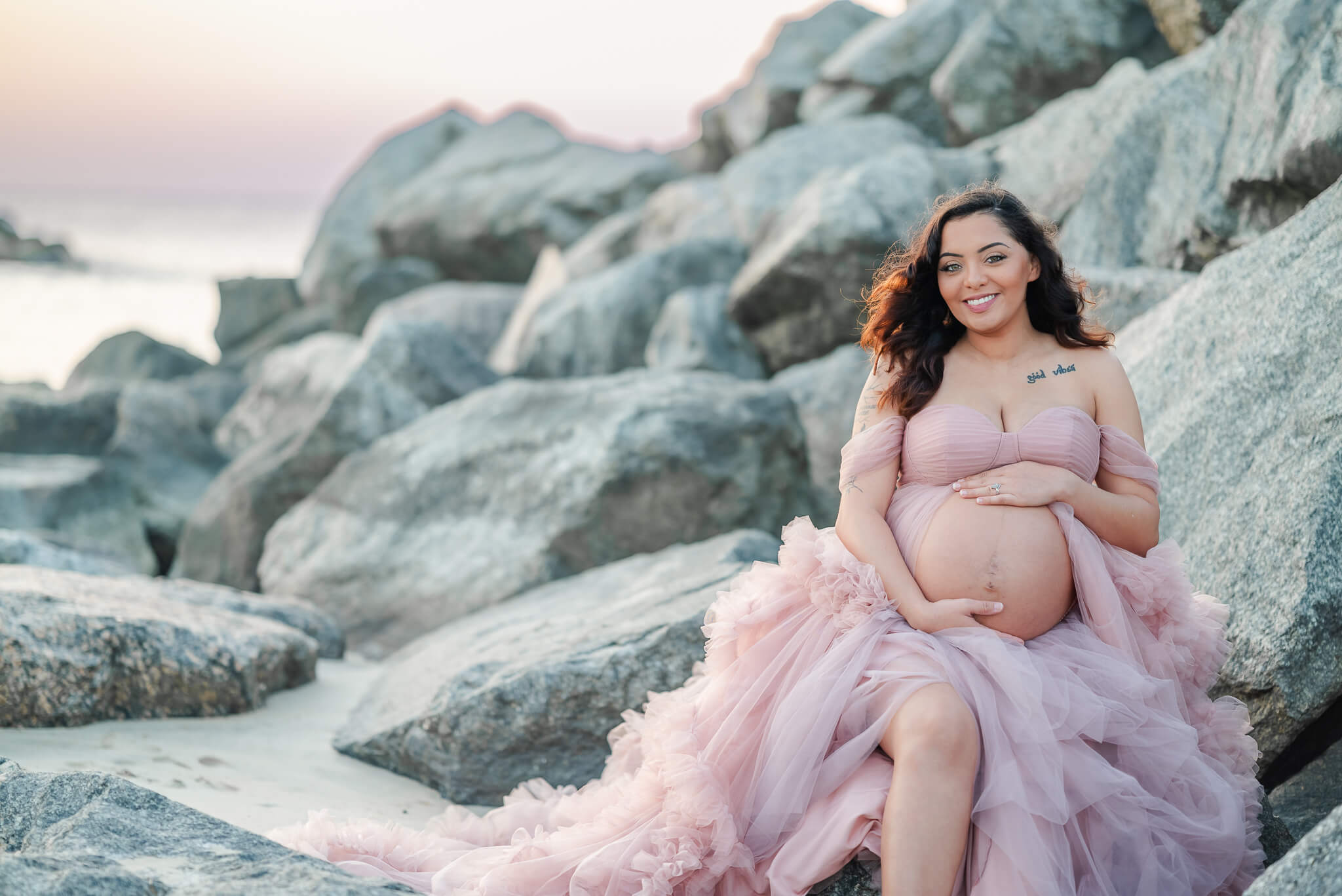 A mama-to-be sits on the rocks on the beach after a relaxing prenatal massage in Chesapeake VA. She wears a pink tulle gown.