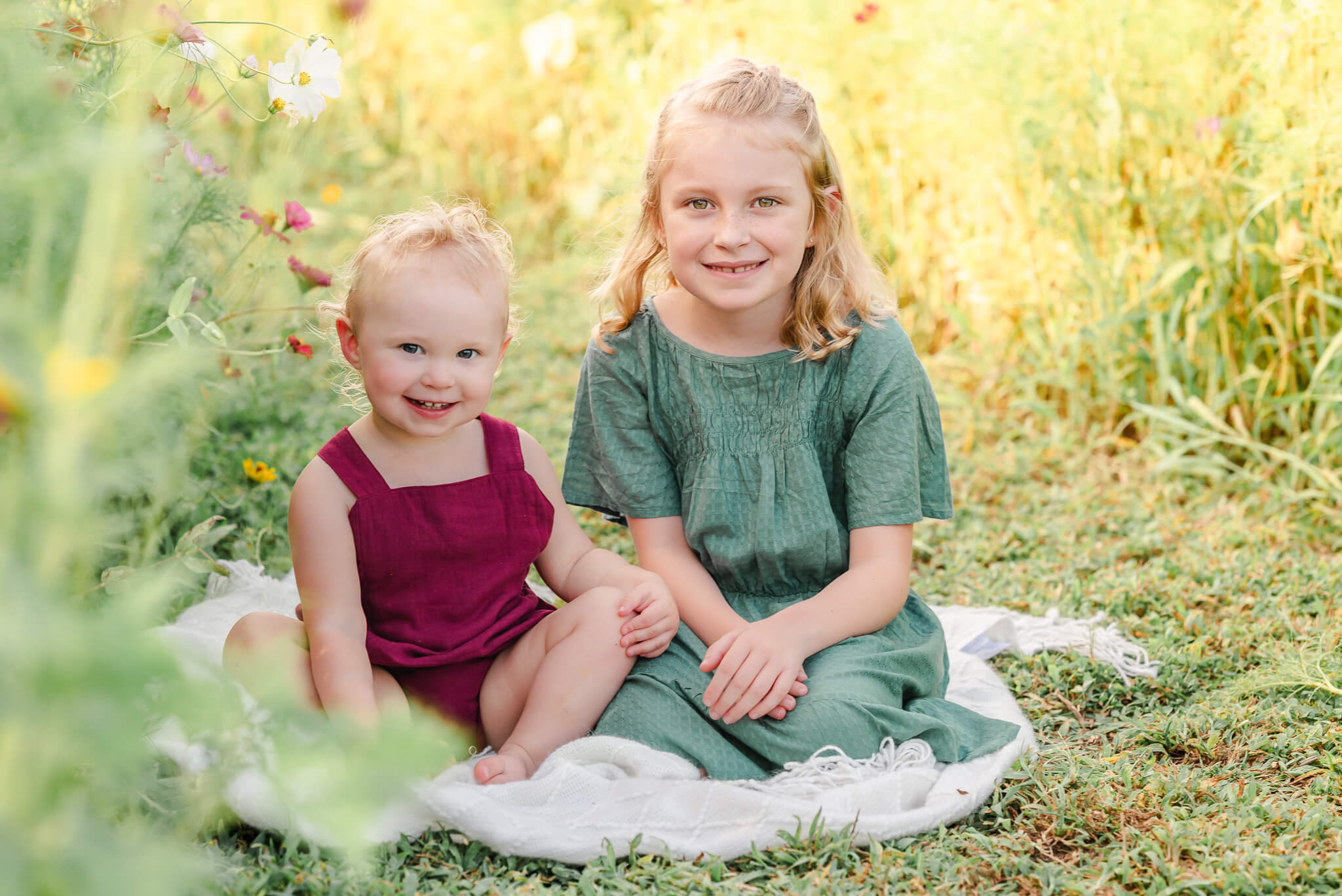 A pair of sisters sits on a white blanket in a field of wildflowers. They wear red and teal. The oldest is old enough to participate in Chesapeake youth sports.