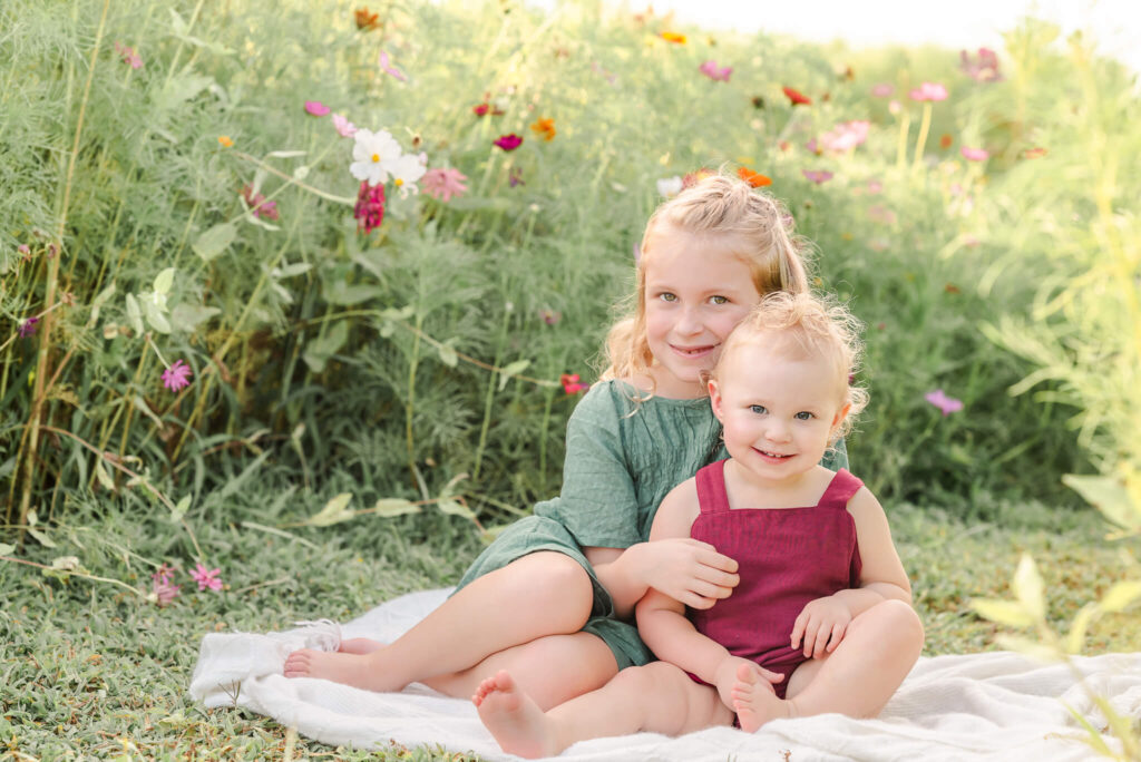 A pair of sisters sits on a white blanket in a field of wildflowers. The youngest wears red and the oldest is in teal. The oldest is old enough to participate in Chesapeake youth sports.