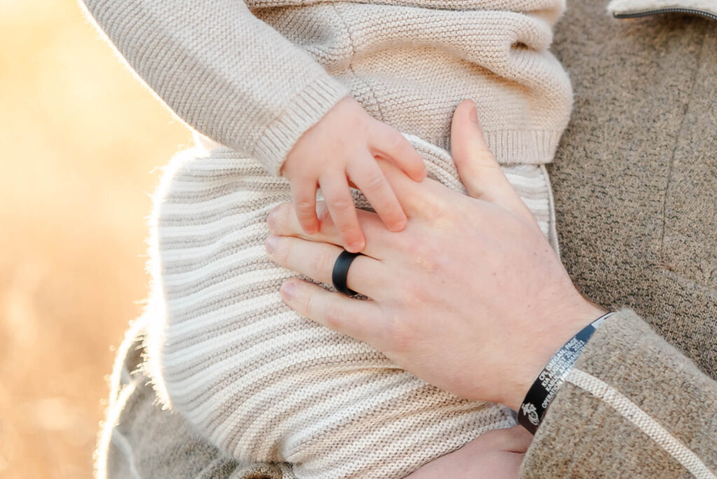 A close up of a toddler holding his dad's hand. The toddler is wear a neutral top and striped pants. This outfit could easily have been found at Little Peas Children's Boutique.