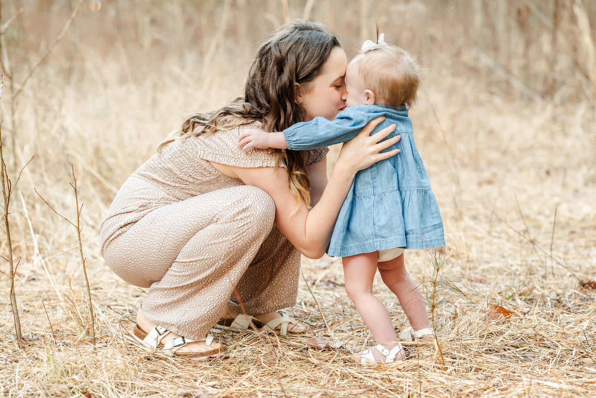 A mom does nose kisses with her toddler daughter. The mom wears a tan jumpsuit and the girl is in a denim dress. The dress could easily have been purchased at Once Upon a Child in Chesapeake.