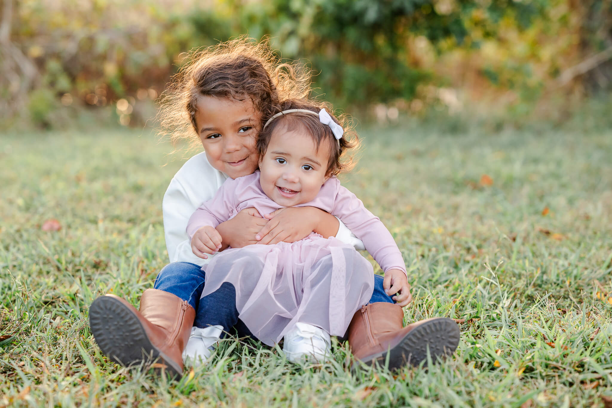 Two toddlers, who participate in preschool in Chesapeake, VA, sit on the grass. The younger one, who is wearing a purple dress, sits on her sister's lap.