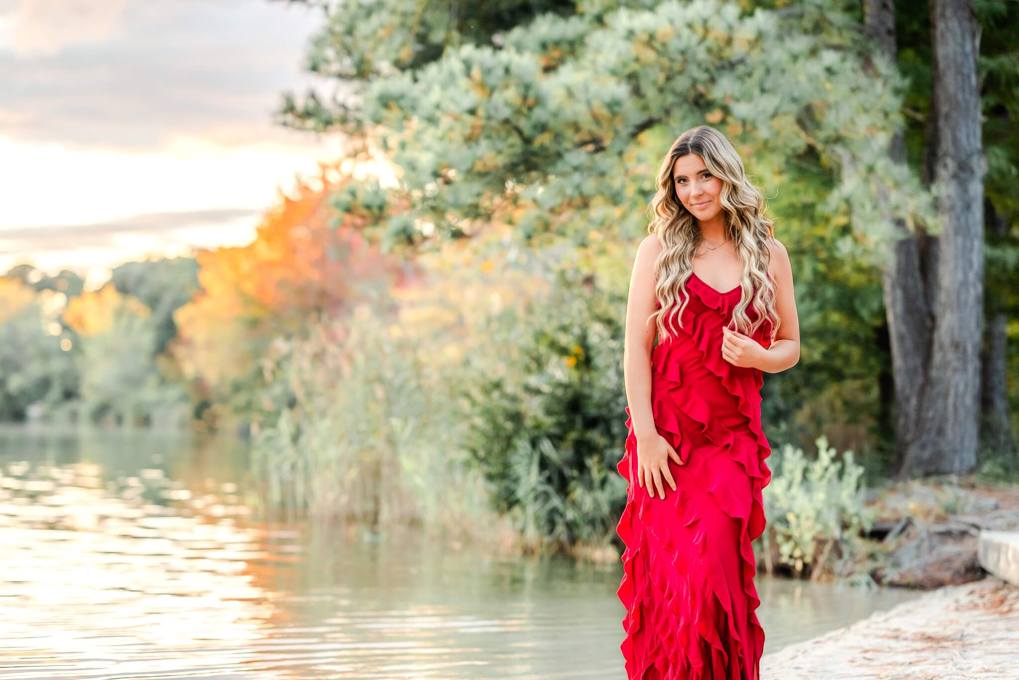 A high school senior stands by a lake at sunset. The glowing sky shows off the just turning fall leaves. She wears a red dress that could be found at All the Rage.