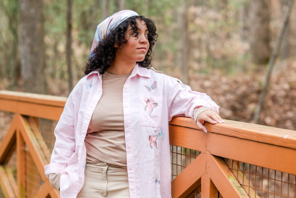 A high school senior wears a pink shirt over a tan undershirt and tan pants. She has a pink bandana in her curly hair. She is leaning on a bridge and looking off into the distance.