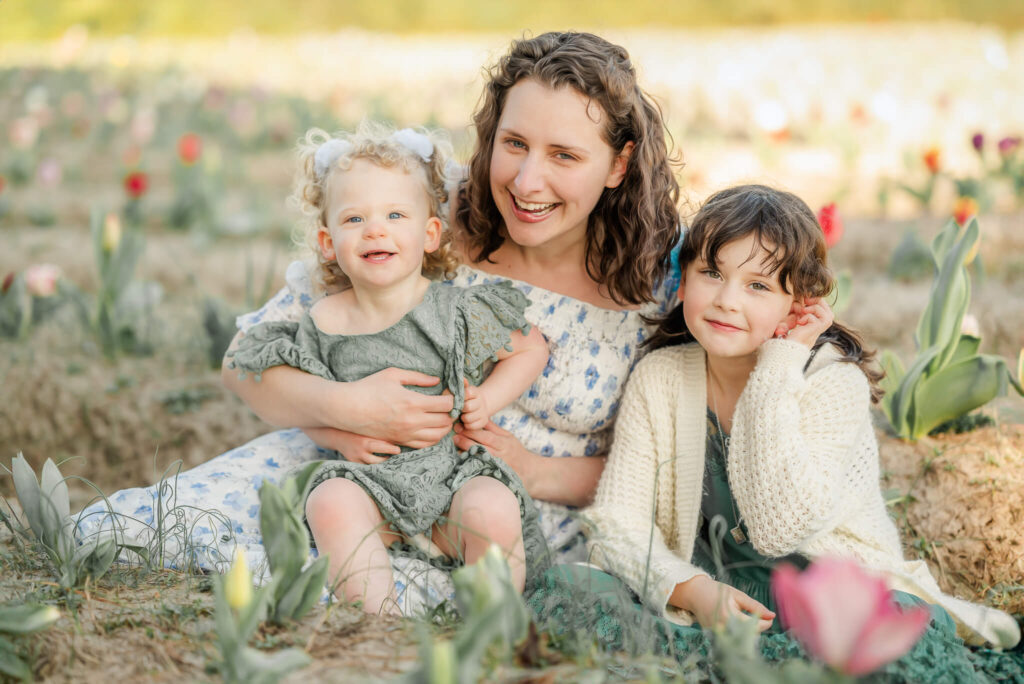 A mom, in a white dress with blue flowers, snuggles in a tulip field with her two daughters, who are both wearing green dresses.