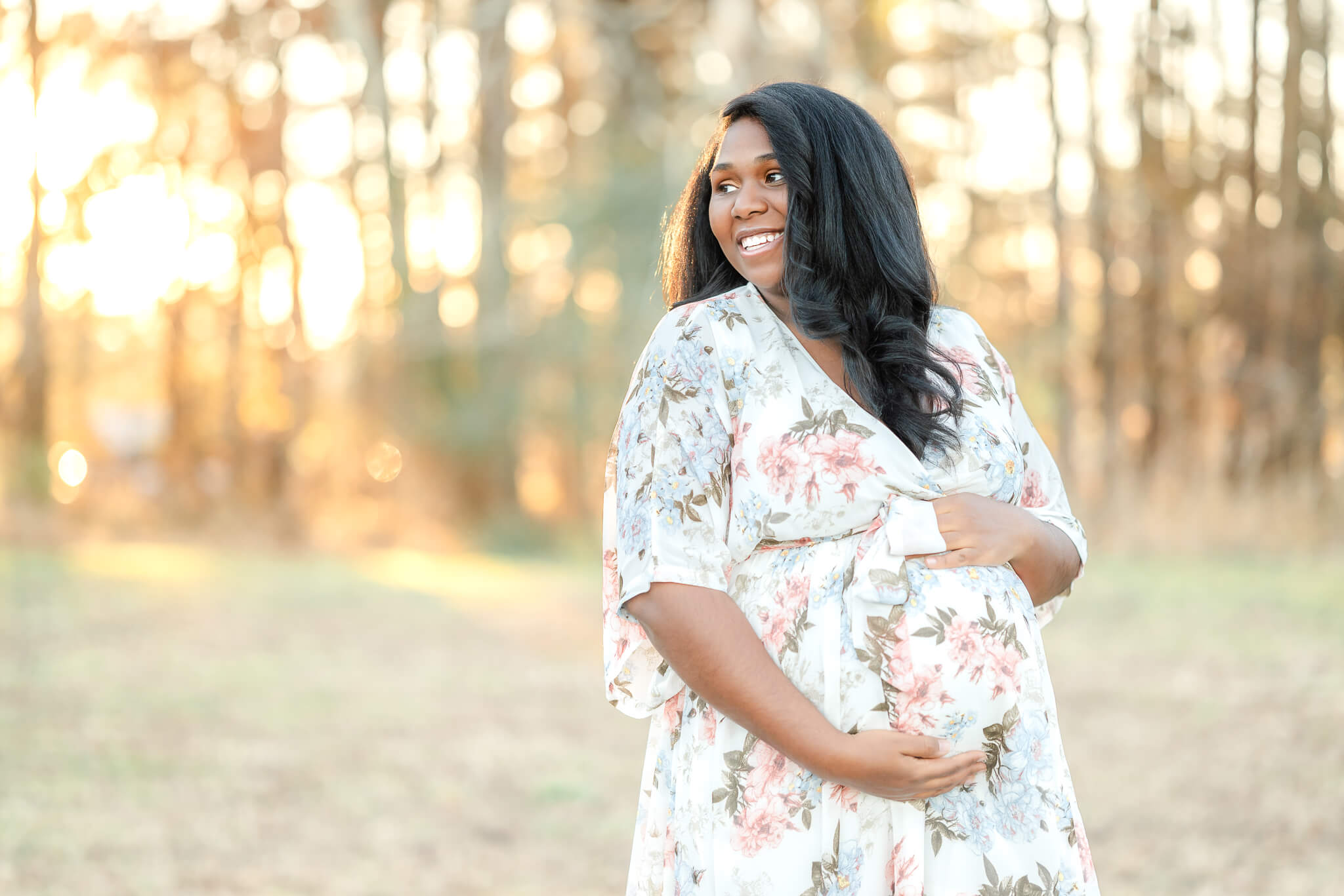A pregnant mama holds her belly and smiles to the side. She is wearing a white floral dress that could have been purchased at The Diaper Bag in Chesapeake.