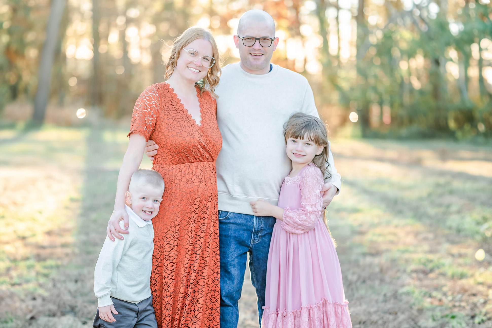 A family of 4 poses for a picture. Dad and son are wearing jeans and off-white sweaters. Mom is in and orange dress and daughter in a pink dress. After the session they went to eat at a family friendly restaurant in Virginia Beach.