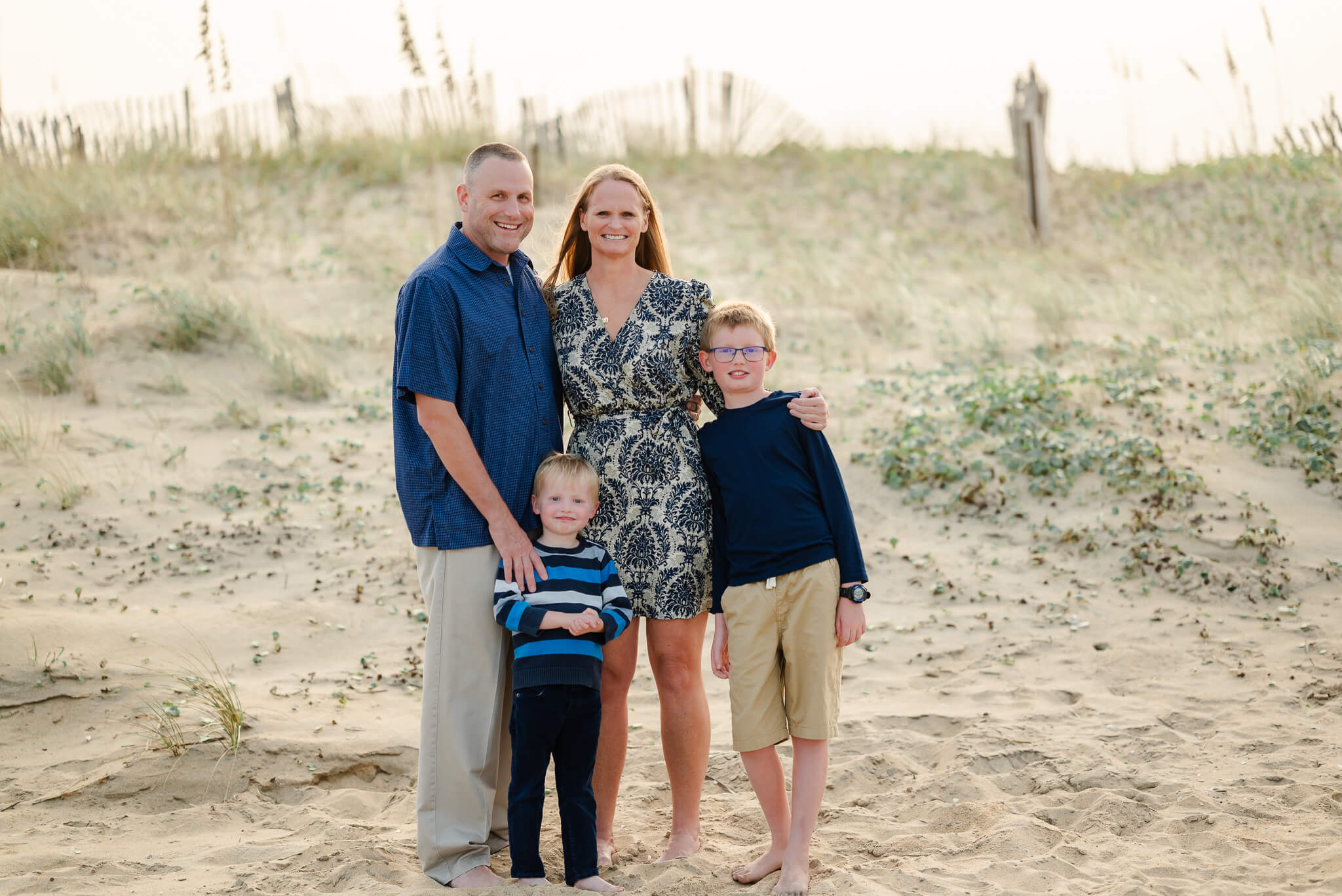 A family wearing blue and khaki stands near the dunes on a beach. They are staying at a kid friendly hotel in Virginia Beach.