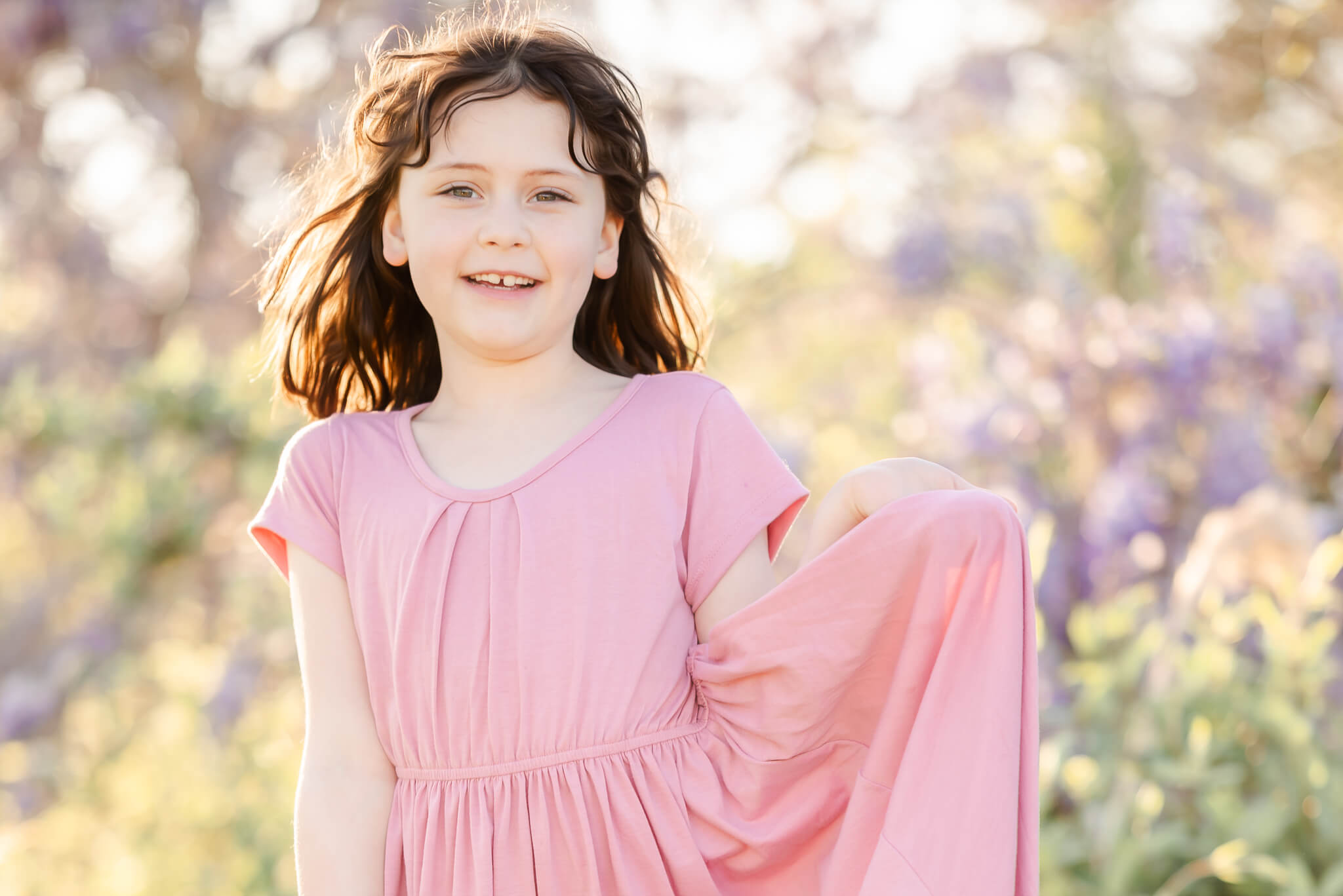A young girl with shoulder length brown hair and bangs wears a long pink dress. She is standing in front of some wisteria. She is the right age for school age classes at the Little Gym in Chesapeake.