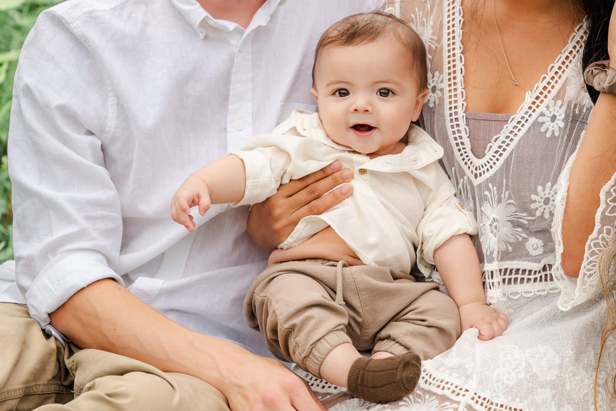 A baby wearing tan pants and an off-white shirt sits on his parents laps. They are in khaki and white. The baby would be in the infant program at Primrose in Chesapeake.
