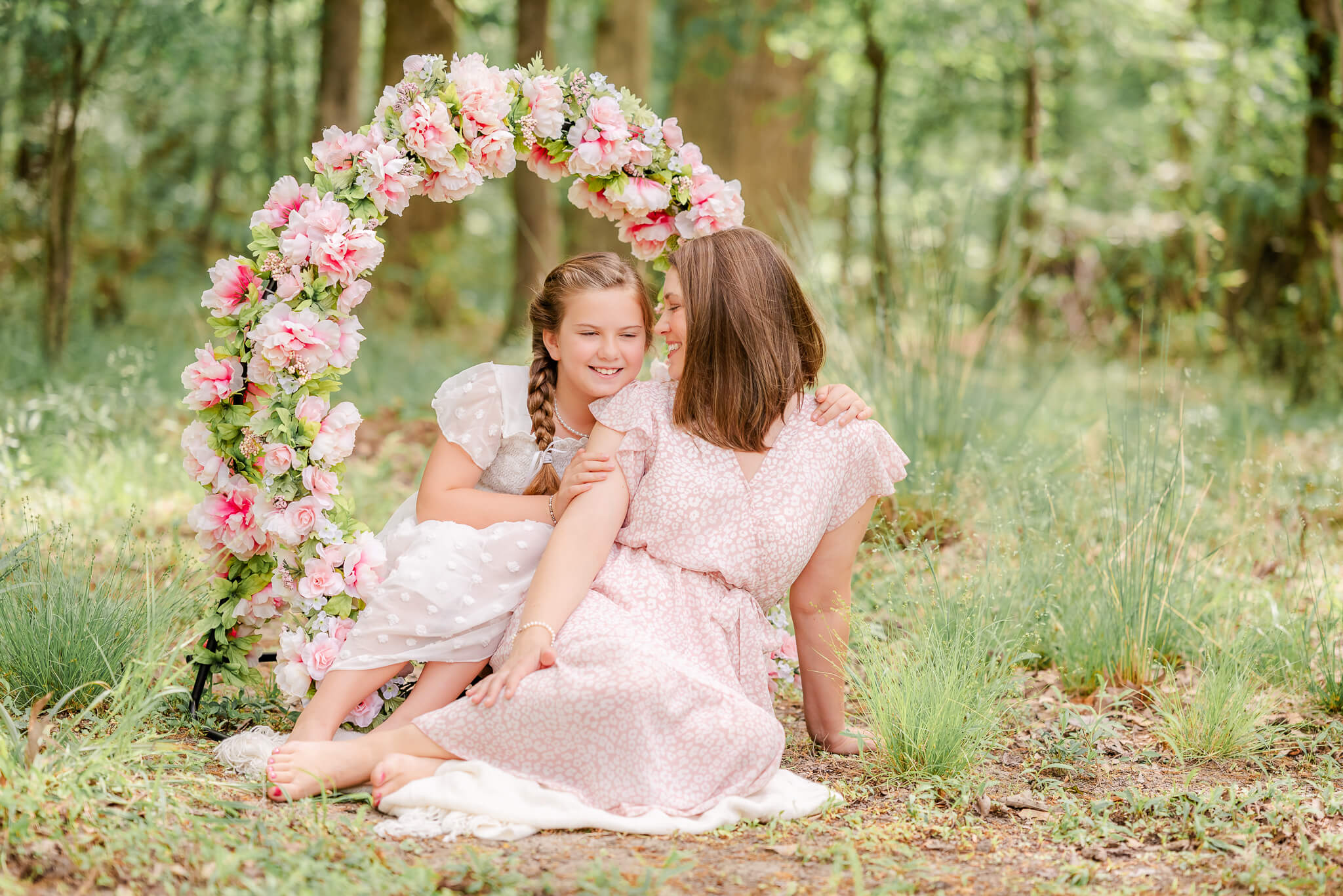 A mother and daughter snuggle close while sitting in front of a floral hoop. The mother wears a pink dress and the girl is in white.