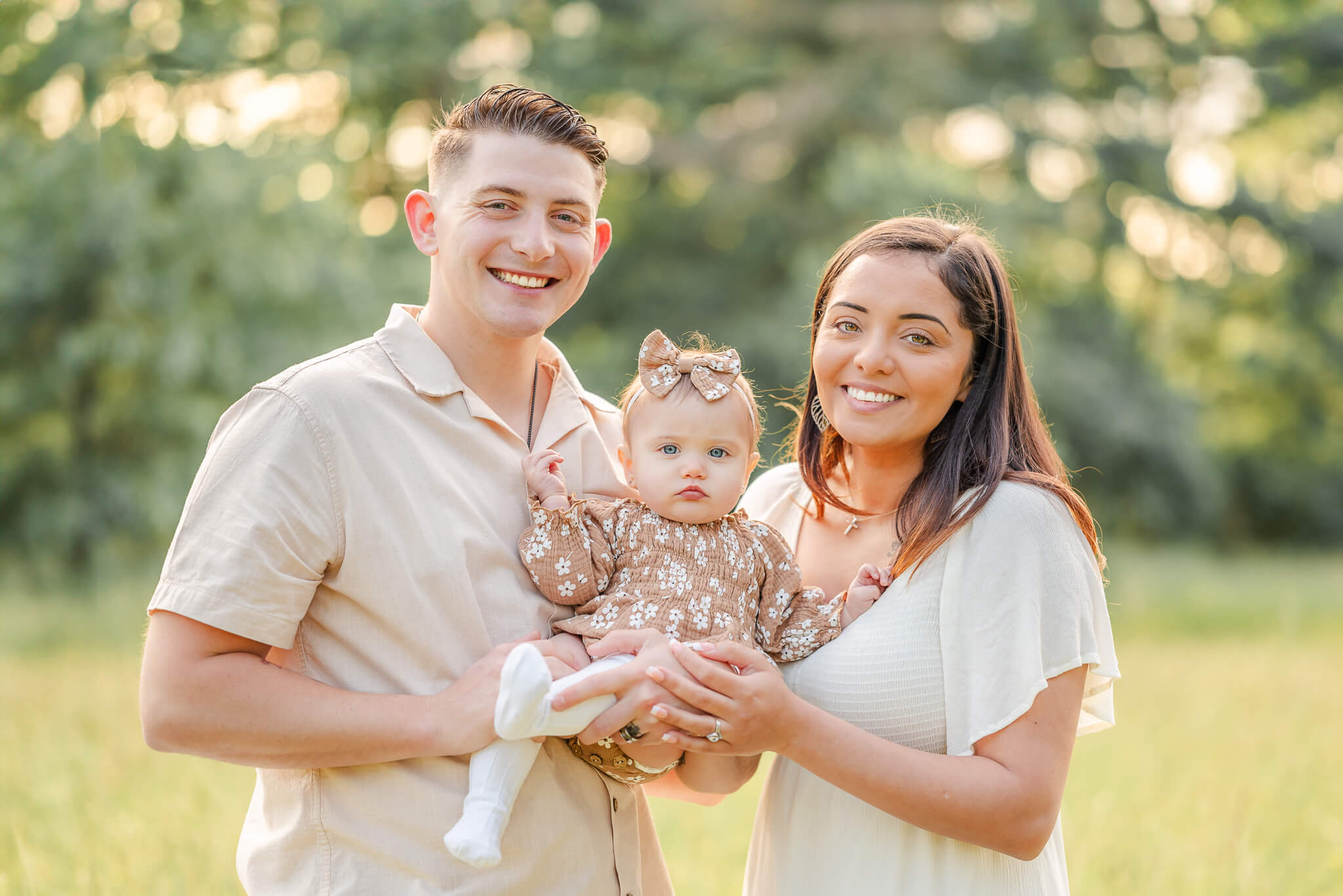 A family of 3 poses in a grassy field. They are dressed in neutrals. The baby, who goes do a daycare in Virginia Beach, wears a brown romper with matching bow.