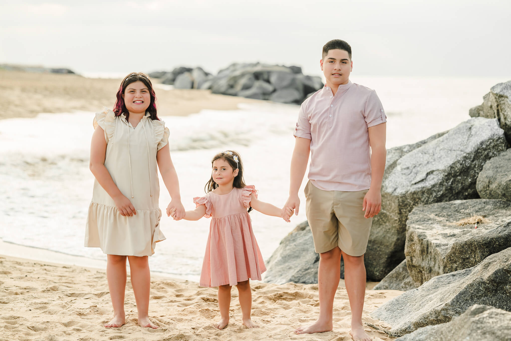 A set of siblings holds hands as they stand in front of the water at Fort Story beach in Virginia Beach. The oldest girl wears and off-white dress. The little girl is in a pink dress and flower crown. The older brother wears khaki shorts and a pink shirt. All the kids are old enough to participate in karate classes in Virginia Beach.