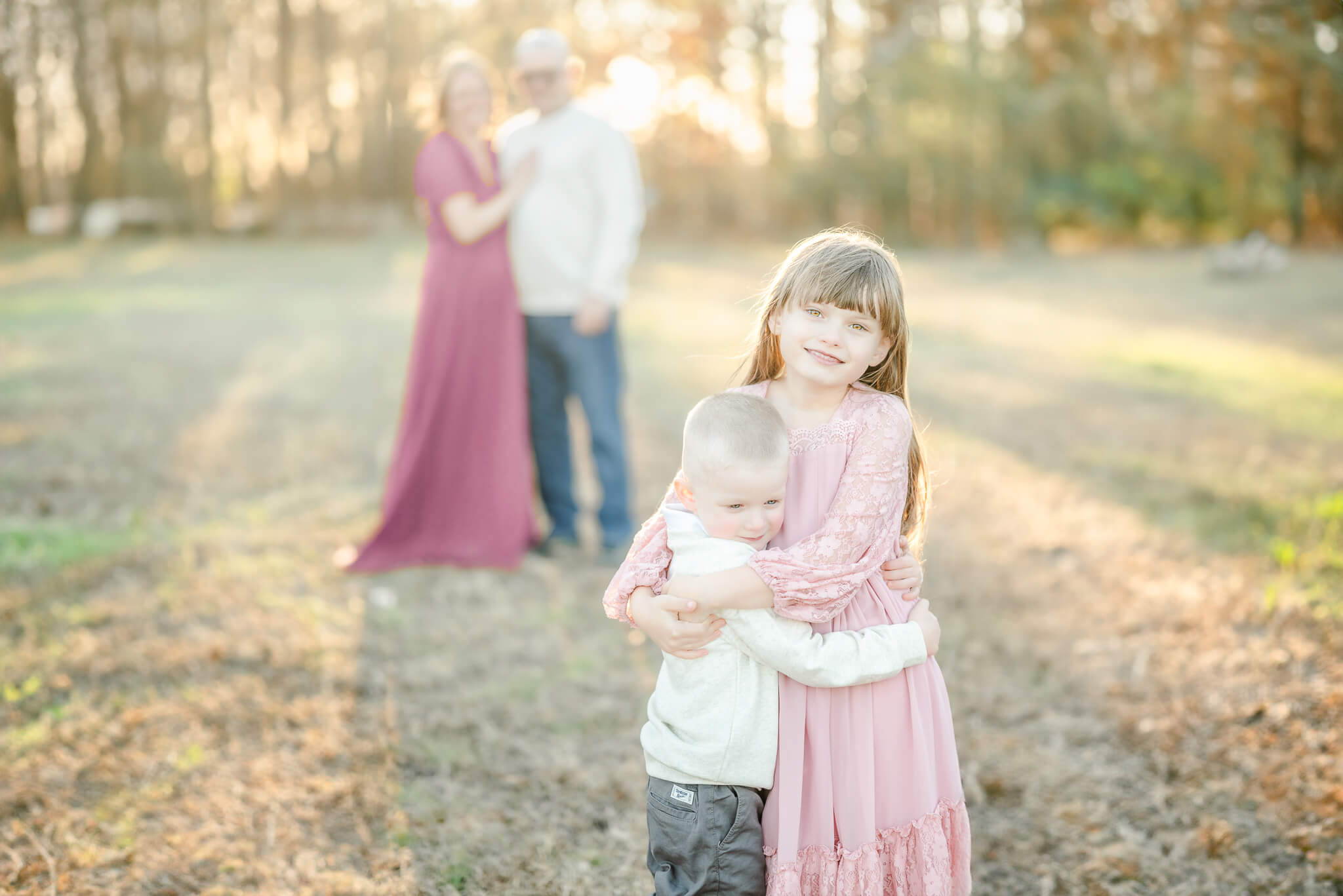 A family of four. The brother and sister are hugging while standing in front of mom and dad, who are in the background. Mom and daughter wear long pink dresses while dad and son are in jeans and off-white sweaters. The children are the right age to attend Montessori school in Virginia Beach.