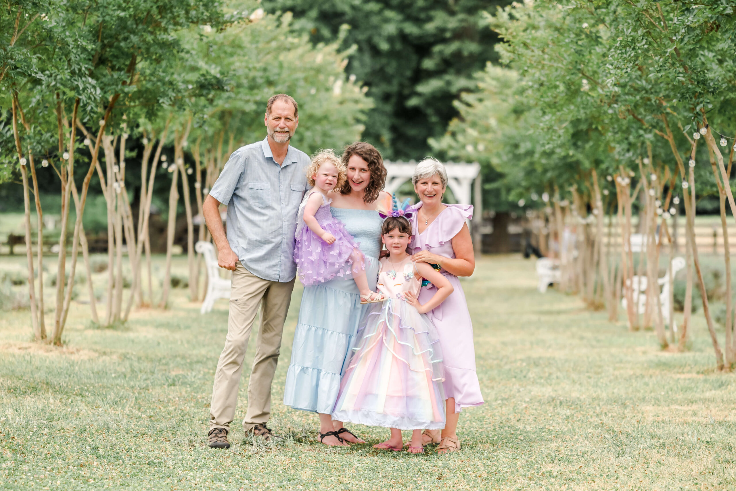 A family stands together between two rows of trees. You can see the grandparents, mom, and two daughters. All are dressed in light blue and purple. The mom could look for nannies in Virginia Beach for her girls.
