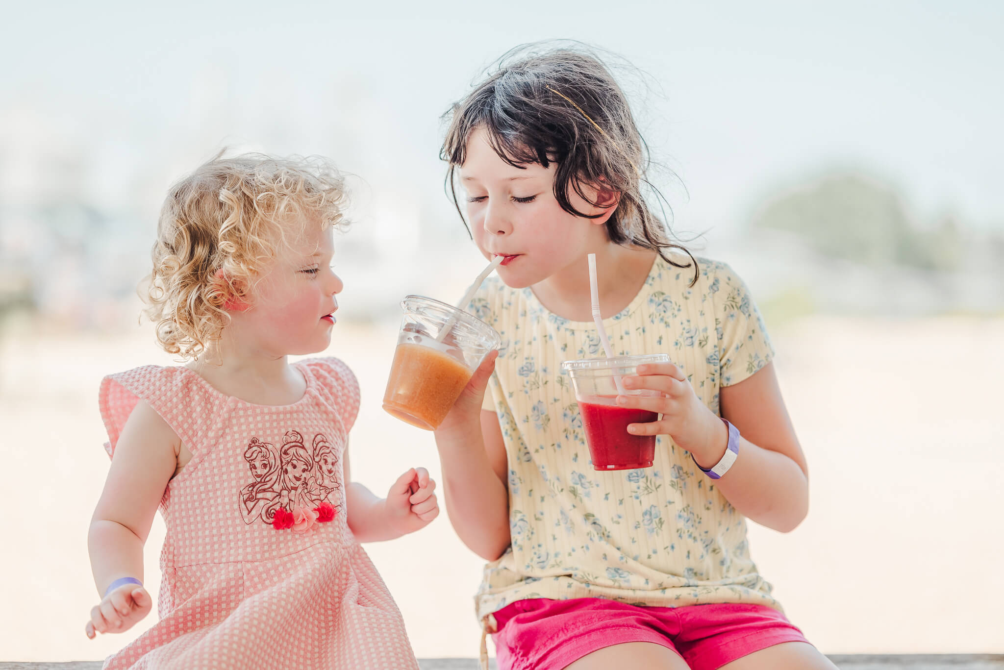 Two sisters enjoy slushies together while sitting on a bench. The toddler is in a pink dress and that child is in a yellow shirt and pink pants. Both see a pediatric dentist in Virginia Beach.