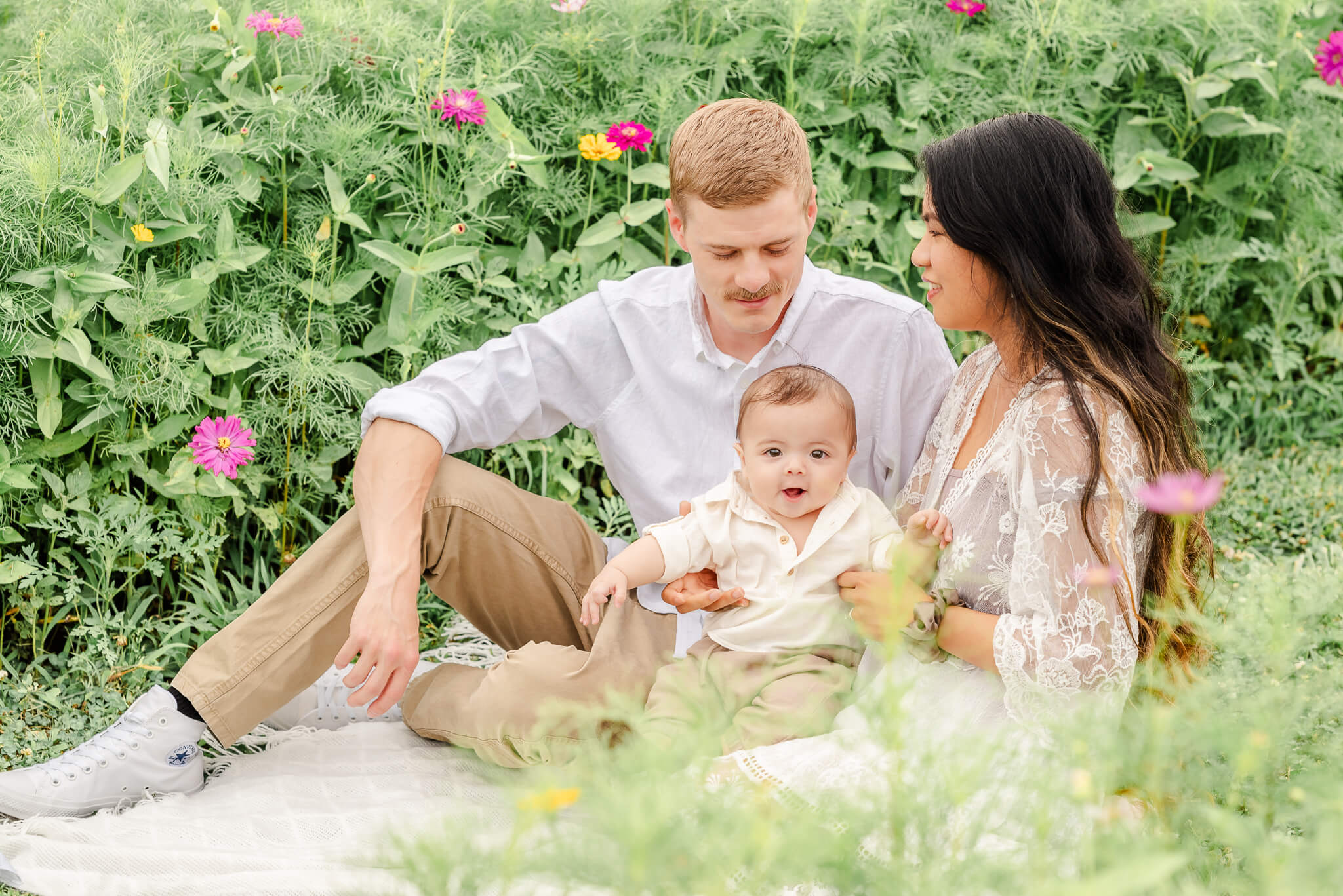 A young family sits in a field of wildflowers. They are dressed in tans and whites. The baby boy sees a pediatrician in Virginia Beach.