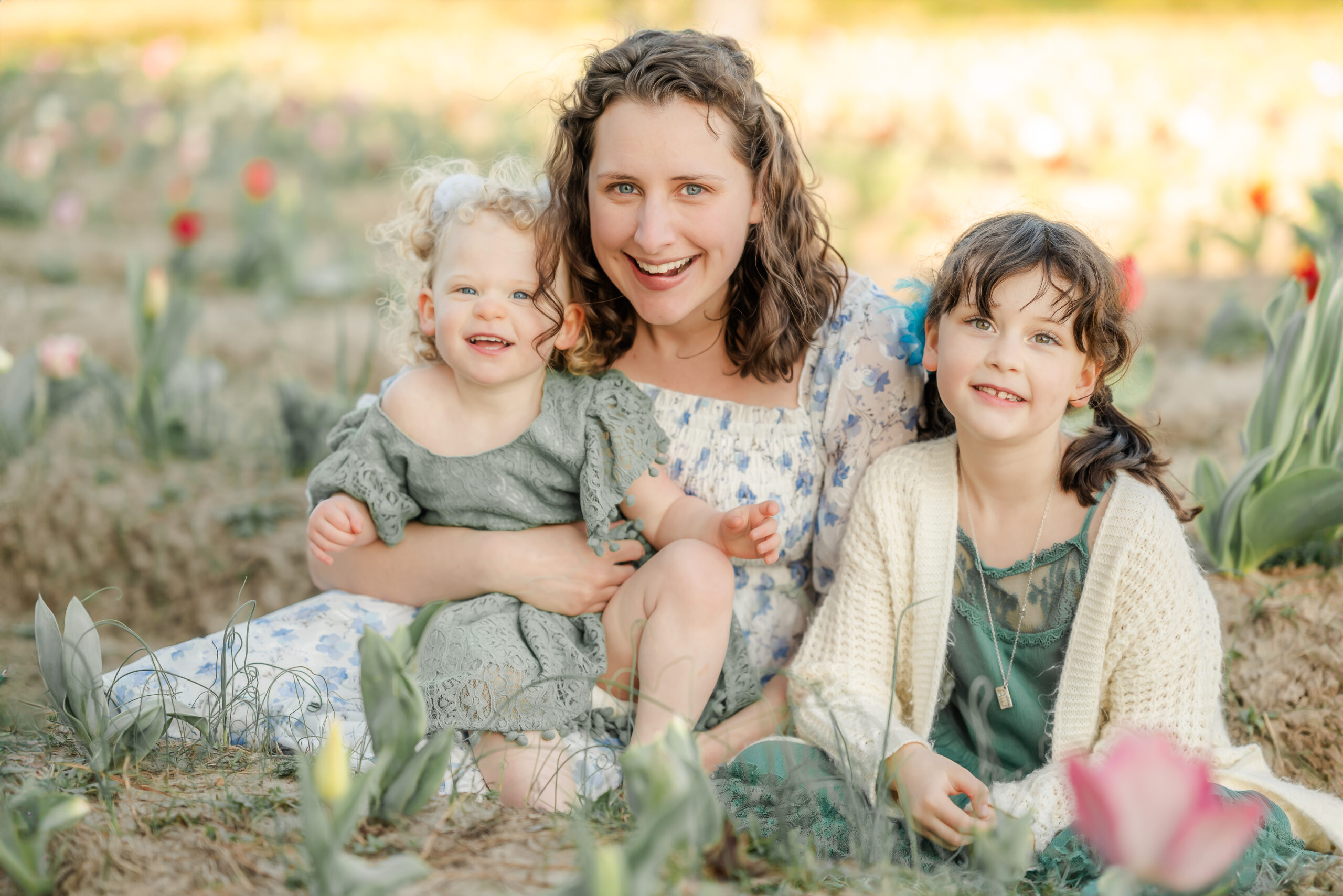 A mother sits in a field of tulips with her two young daughters. They are wearing greens and whites. Flower picking is an example of a family activity in Virginia Beach.