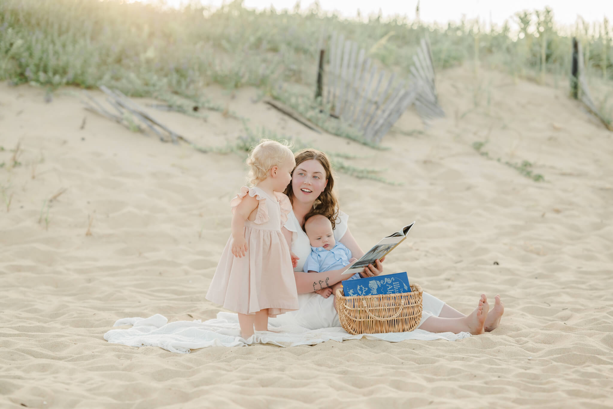 A mother sits on a white blanket on the beach and reads to her baby son and toddler daughter. A picnic on the beach is one of the many family-friendly things to do in Virginia Beach.