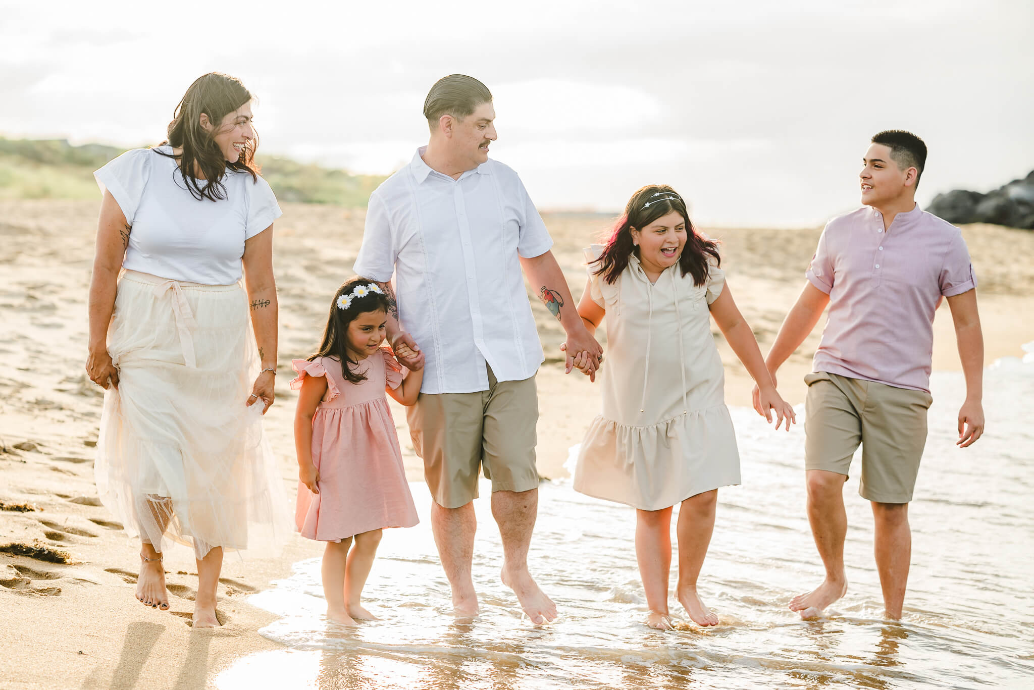 A family of five, dressed in neutrals and pinks, walks along the beach while holding hands. They look at each other and smile because the beach is the perfect place for family fun in Virginia Beach.