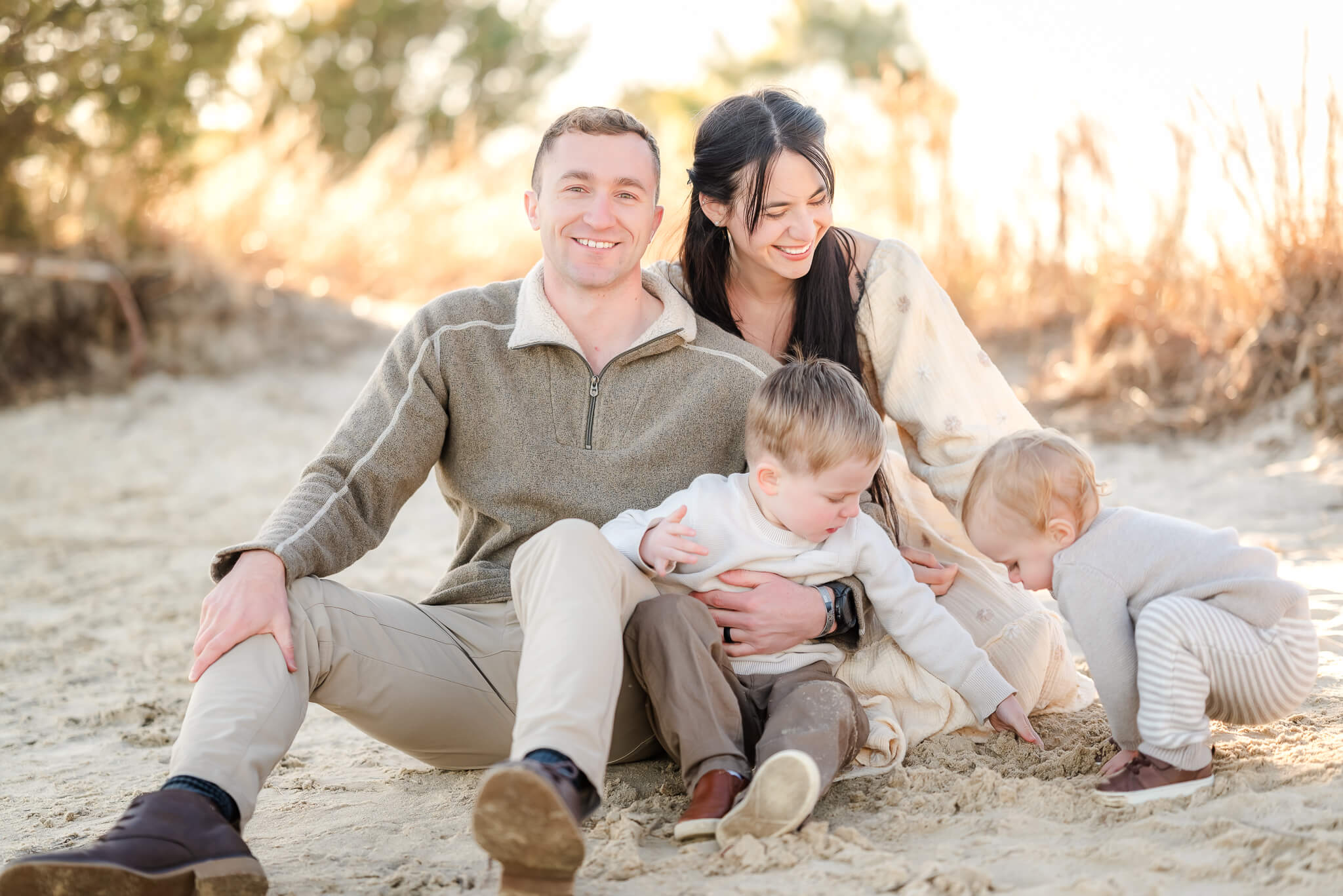 A family of four sits in the sand. They are wearing neutral outfits and the sun is shining behind them. The toddler boys benefit from Virginia Beach childcare while their parents work.