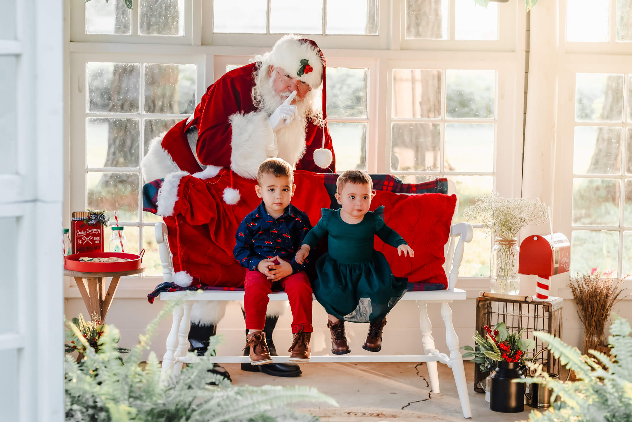 Santa stands behind a bench with his finger to his lips. Two toddlers sit on the bench in front of him. Seeing Santa is a great Virginia Beach holiday event.