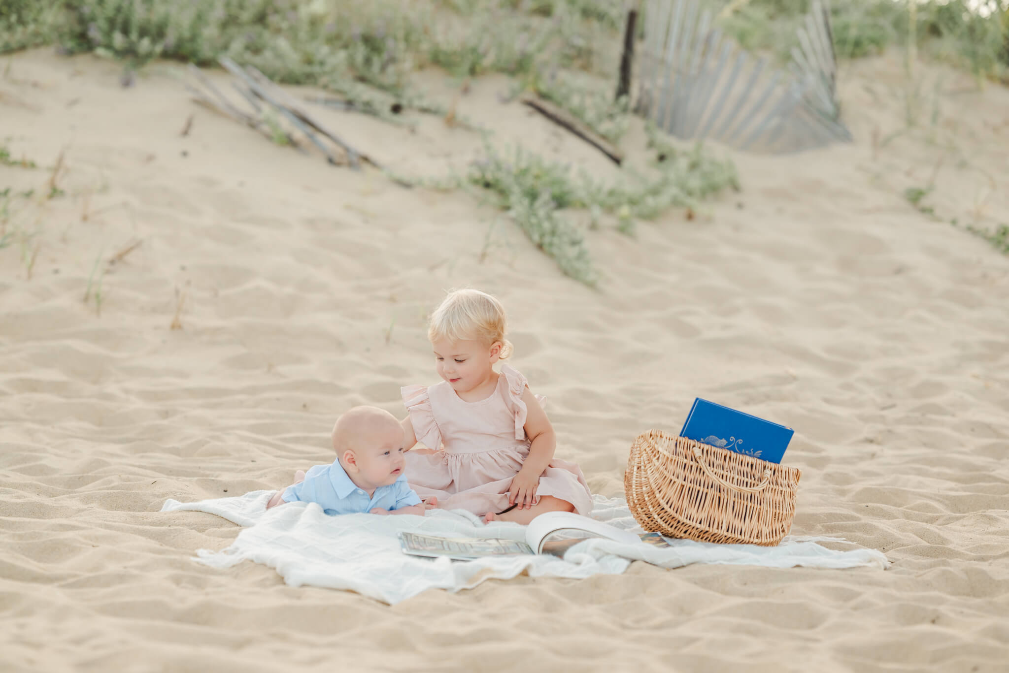 A toddler wearing a pink dress sits on a white blanket on the beach with her baby brother. There is a basket of books by her. She enjoys reading books while attending preschool in Virginia Beach.