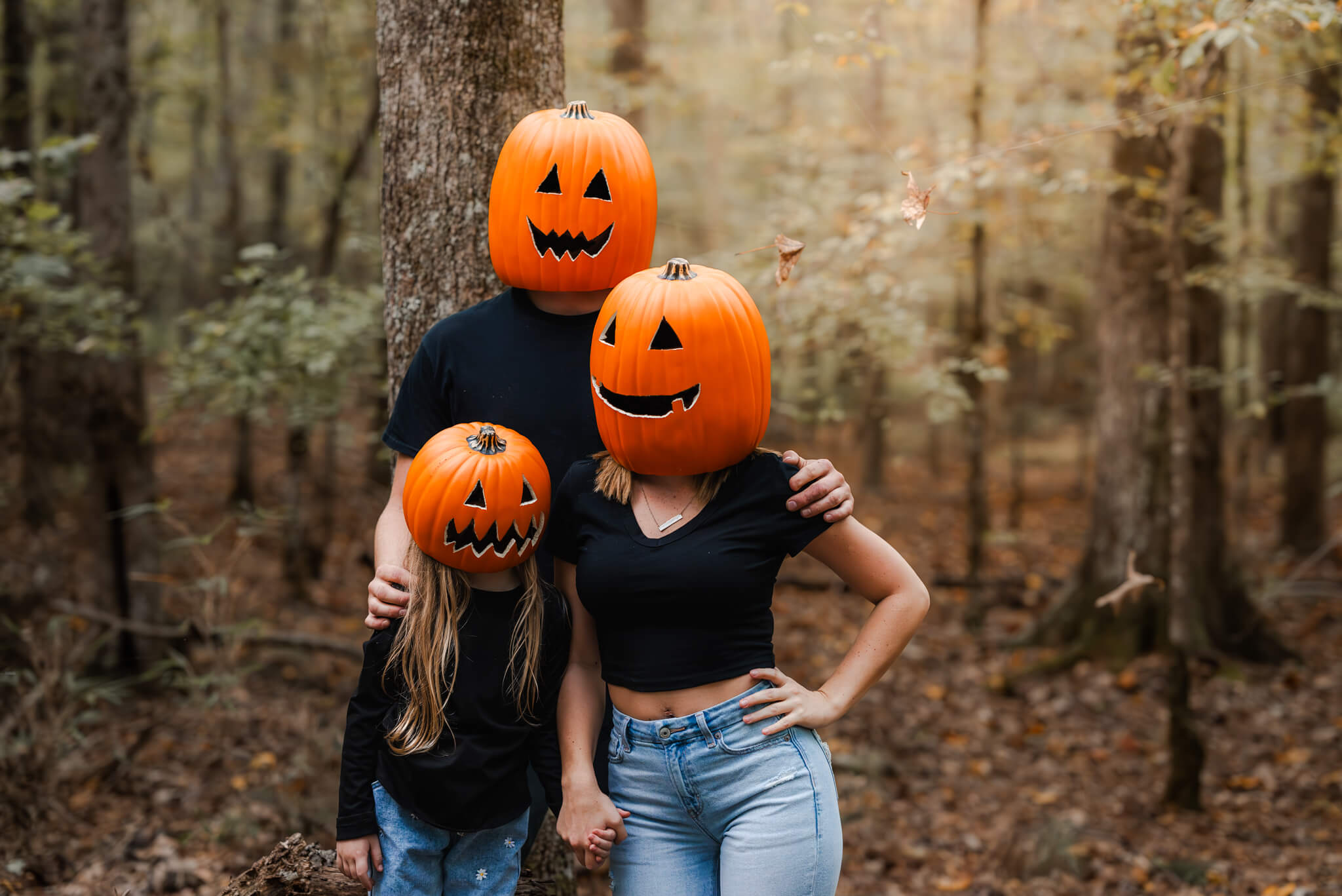 A family of three, wearing black shirts, jeans, and jack o'lantern heads poses for a photo in a forest. Getting spooky portraits is a fun Chesapeake Halloween event.