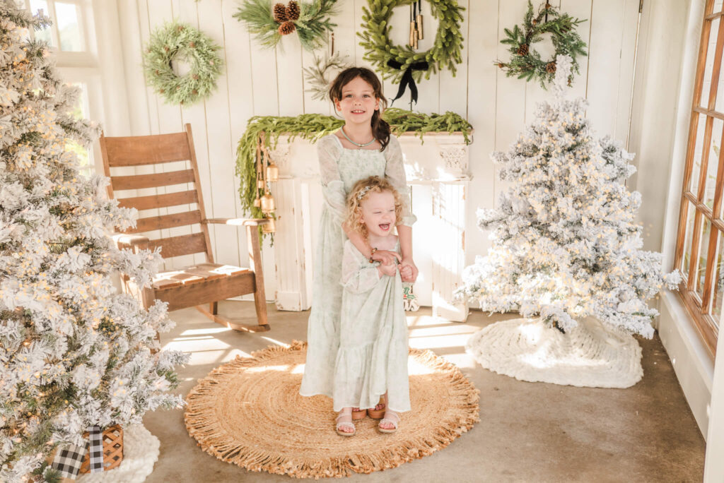 Two young girls in long, white dresses with sage flowers, stand in a sun porch decorated for Christmas.