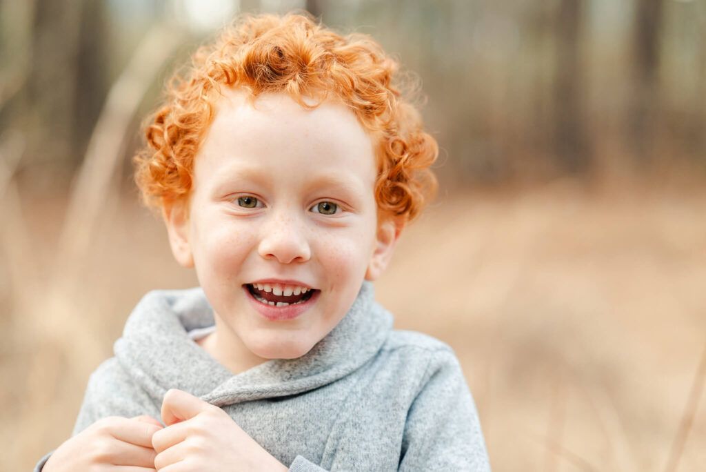 A young red-headed boy smiles big. He is wearing a gray sweater and playing with the button on it.