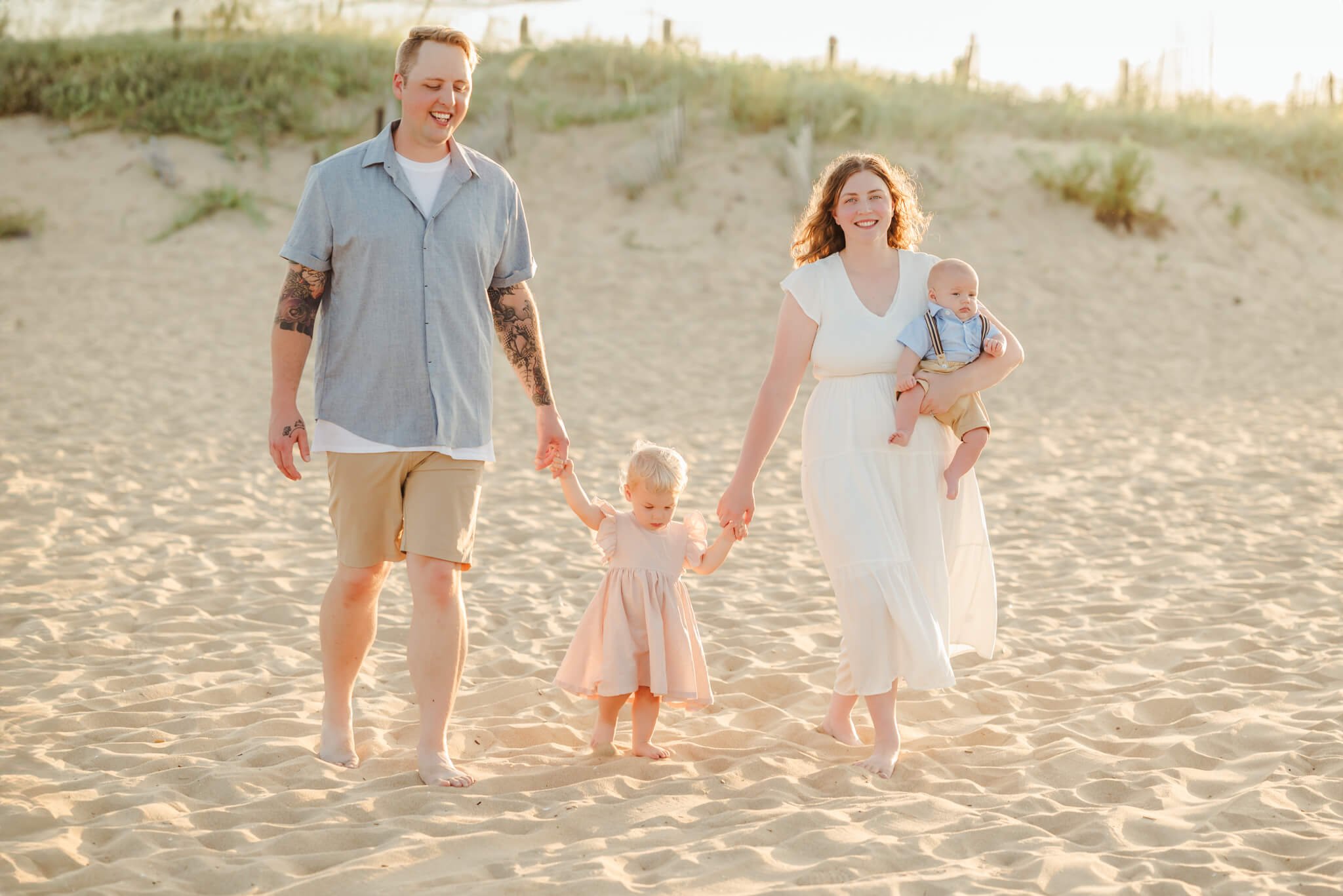 A family holds hands while they walk on the beach. The beach dunes are behind them. The mother has the baby in her arms. They are relaxed and happy. You can clearly see them enjoy their session.