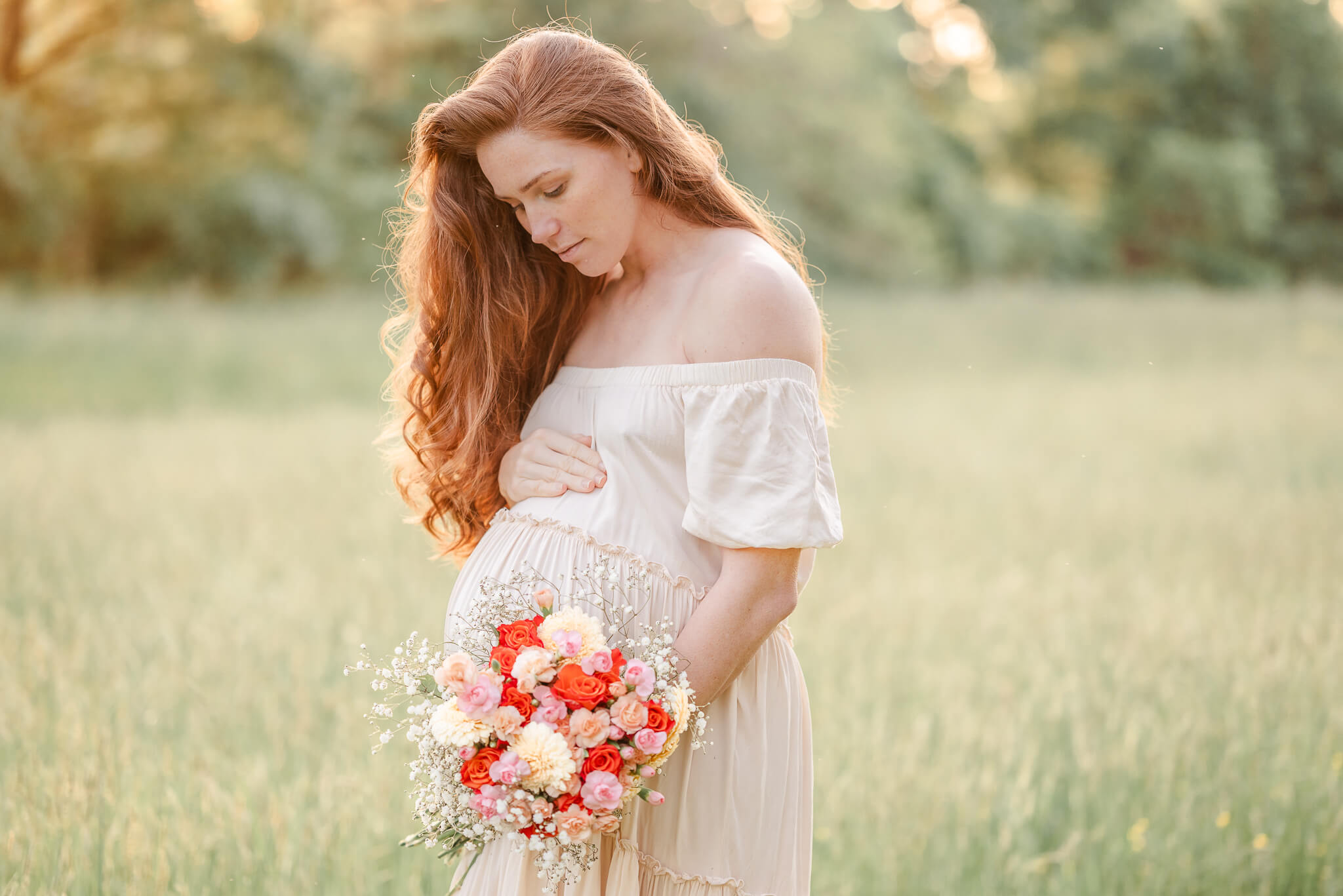 An expectant mother holds a bouquet of flowers and looks down at her bump. The off-white dress she is wearing looks amazing against her red hair and the green grass of the field. She would enjoy the gift of a photo session when her baby comes along.